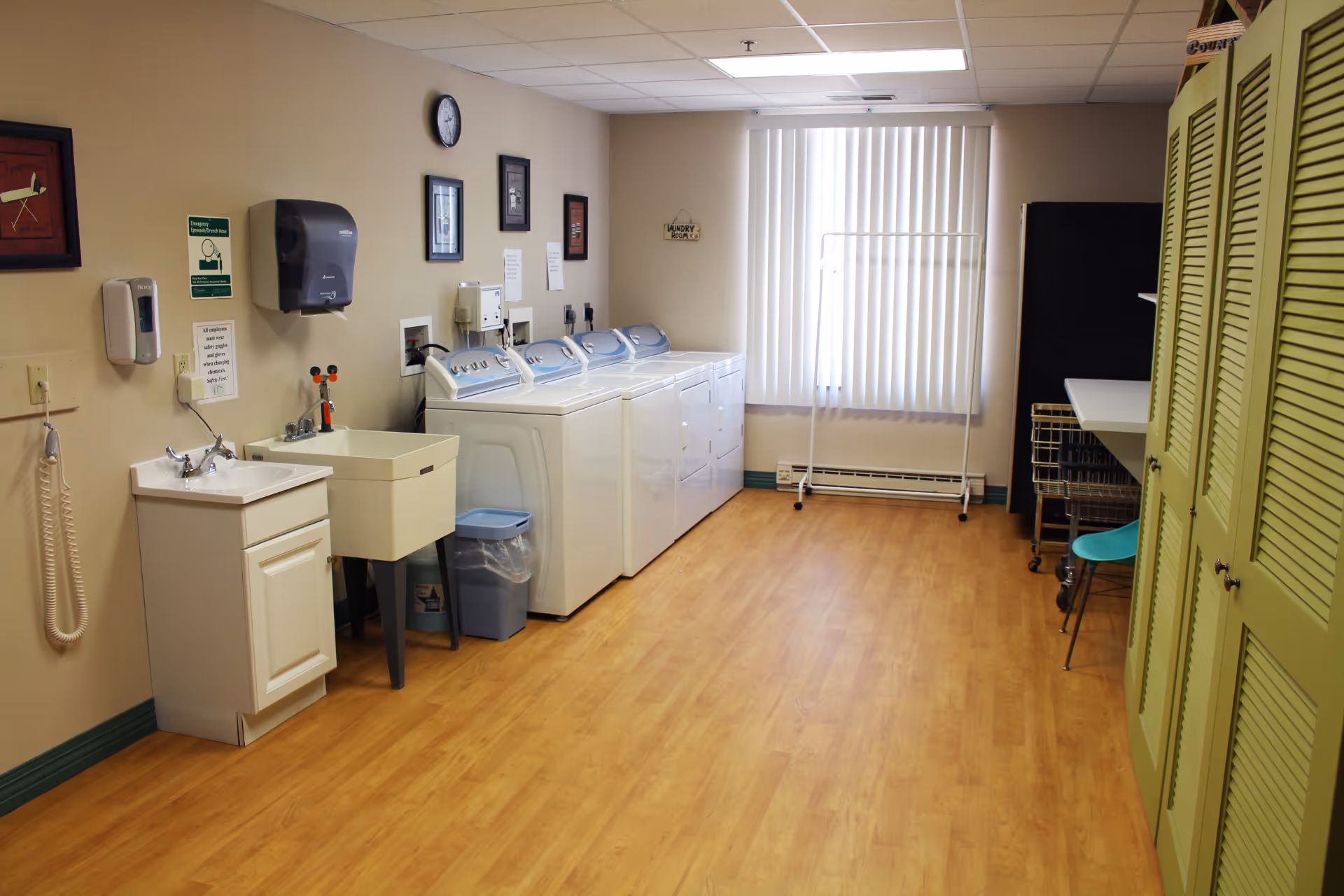 A clean laundry room with multiple washers and dryers, utility sinks, folding table, carts, and green lockers along the wall.