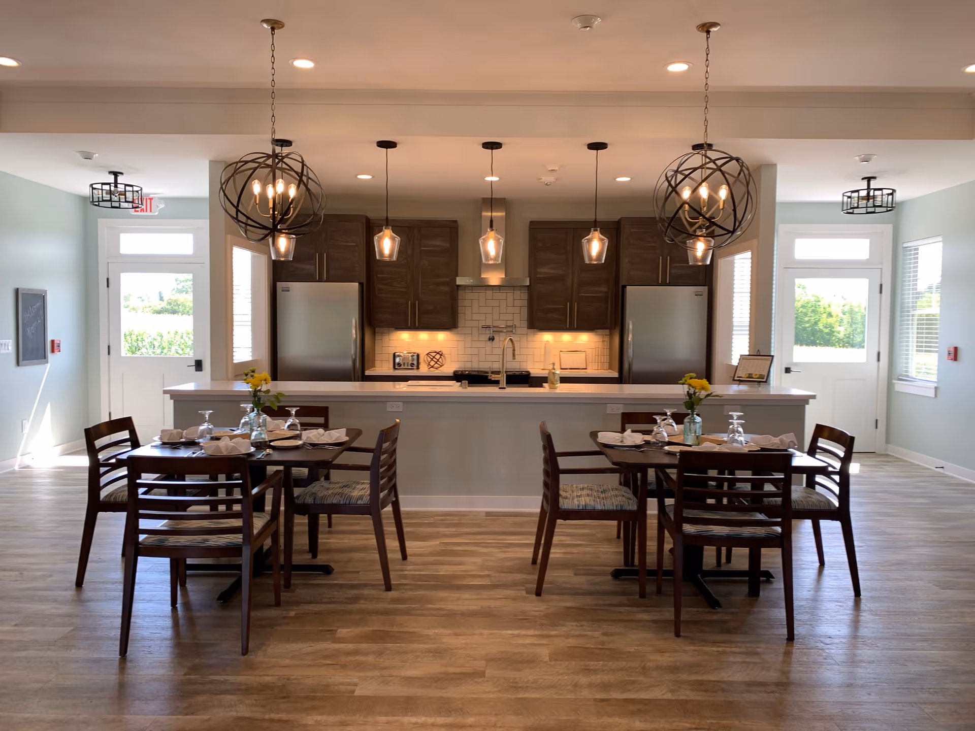Open dining area with two set wooden tables and chairs facing a modern kitchen island with pendant lights and stainless steel appliances.