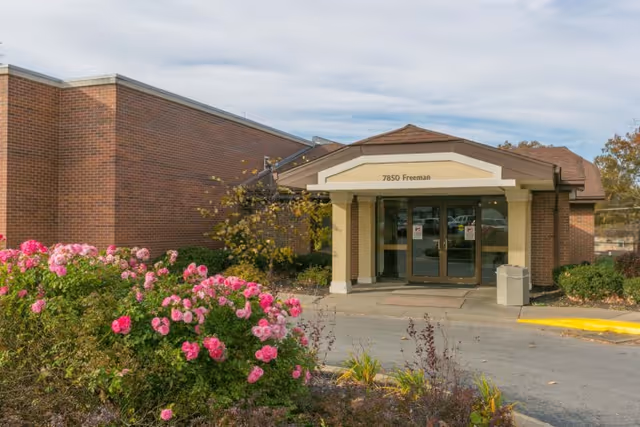 Entrance of a brick building with a covered porch and double glass doors. The address 7850 Freeman is displayed above the entrance. There are pink flowers and green shrubs in the foreground, and the sky is partly cloudy.
