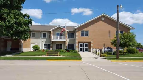 Two-story brick and beige senior living building with entrance, flag, and manicured lawn.