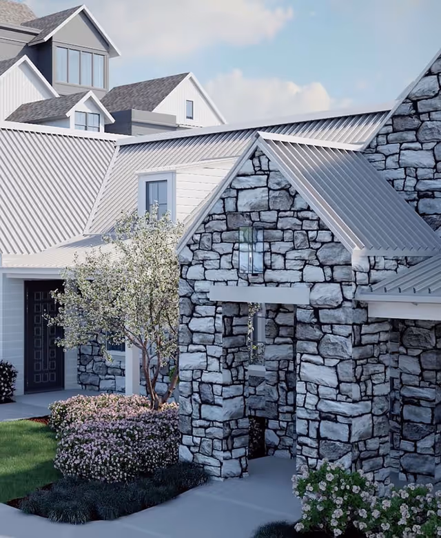 Exterior view of a senior living facility building with stone and white siding walls, metal roofing, a small tree with white blossoms, and landscaped bushes with pink flowers in front.