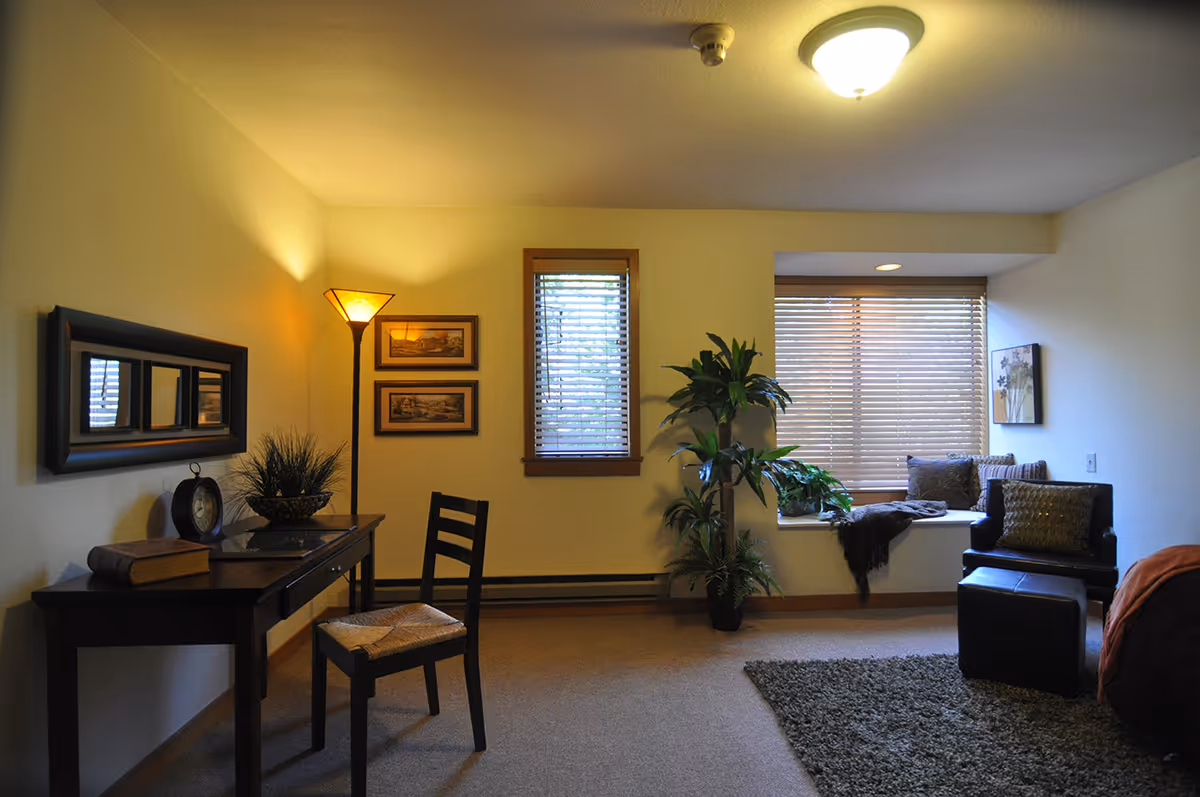 A cozy interior room with a wooden desk and chair on the left, decorated with a table lamp, a clock, a book, and a plant. There are two framed pictures on the wall above the desk and a rectangular mirror. In the center, there is a window with wooden blinds and a tall green plant. On the right side, there is a window seat with cushions and a throw blanket, next to a black armchair and matching ottoman on a gray rug. The room has beige walls and carpeted floor with ceiling light fixtures.