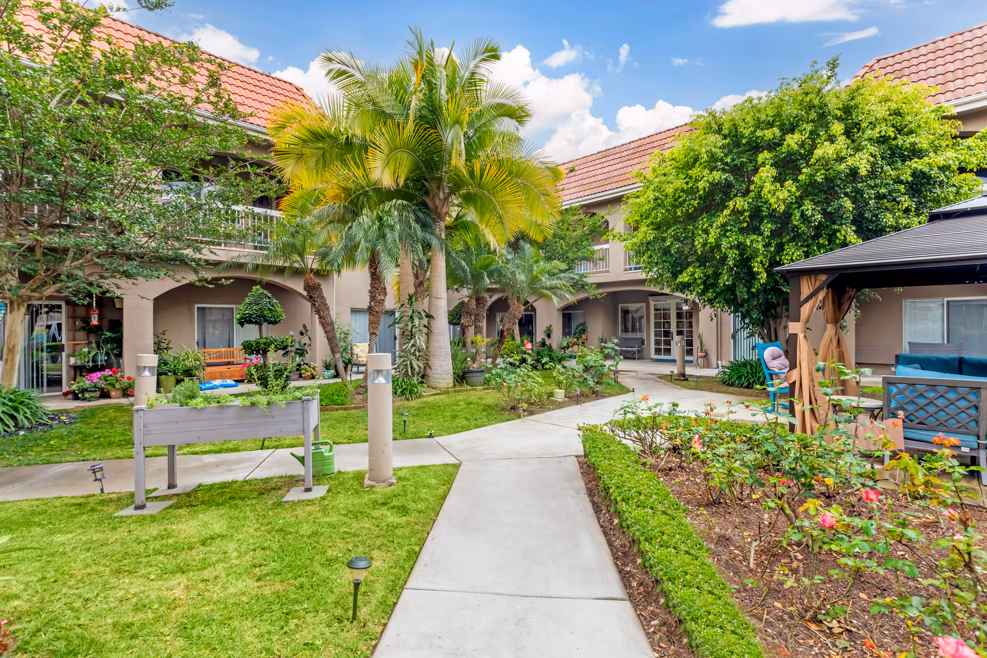Outdoor courtyard area at Brookdale Brea featuring a paved walkway surrounded by green grass, palm trees, flowering plants, and shrubs. There are benches and a gazebo with seating on the right side, and the building with balconies and red-tiled roof is visible in the background under a partly cloudy sky.