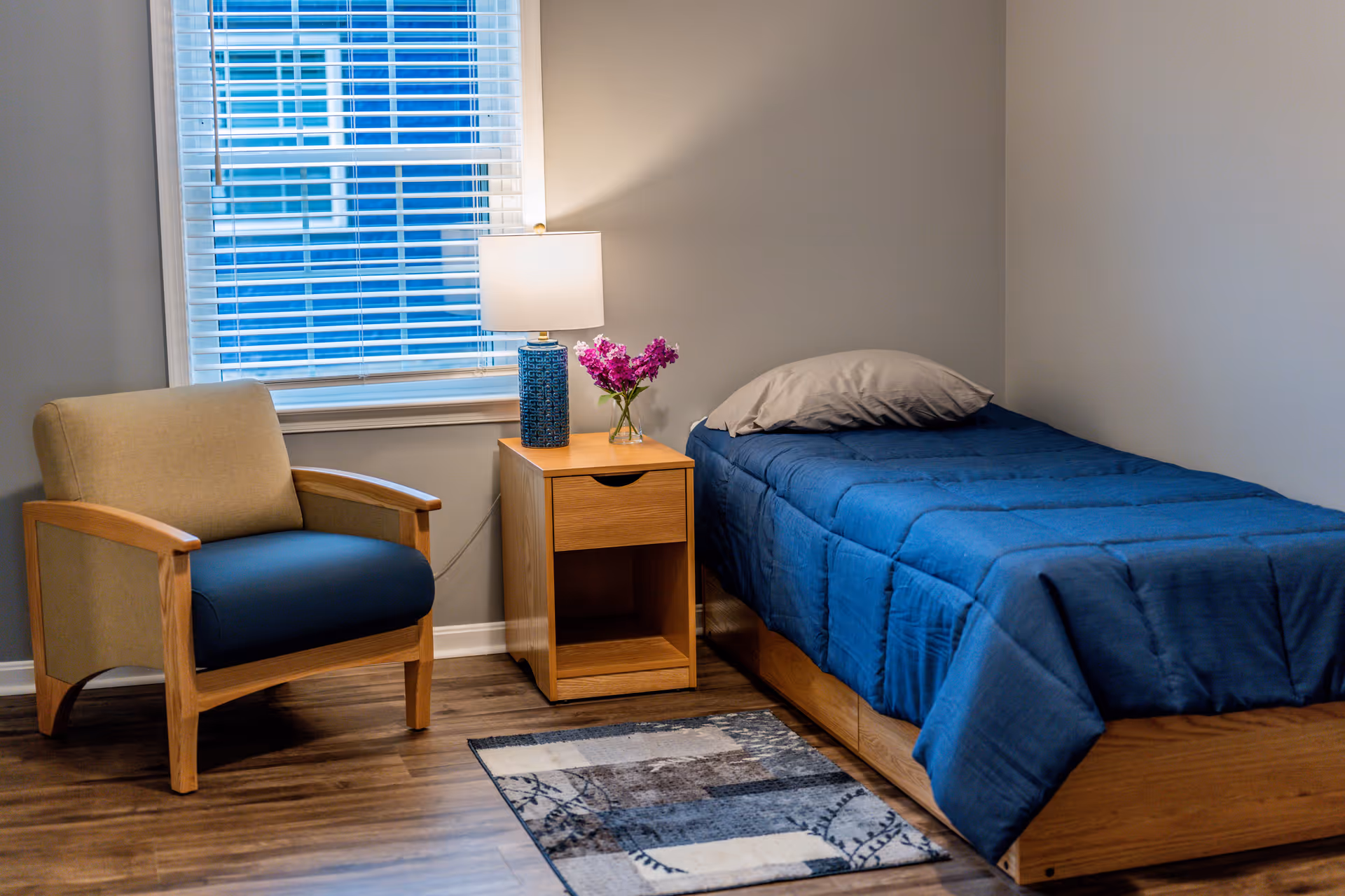 A small bedroom with a single bed covered in a blue comforter and a gray pillow. Next to the bed is a wooden nightstand with a blue lamp and a small vase of purple flowers. A beige and blue armchair is positioned near a window with white blinds. The floor is wooden with a small patterned rug in front of the bed.