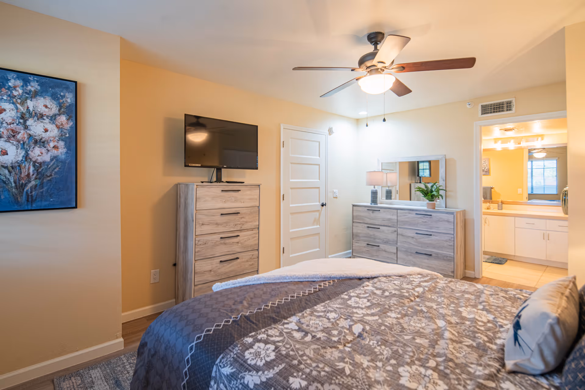 A bedroom with a bed covered in a patterned comforter, a ceiling fan with light, a dresser with a mirror and lamp, a chest of drawers with a flat-screen TV on top, and a doorway leading to a bathroom with a sink and mirror.