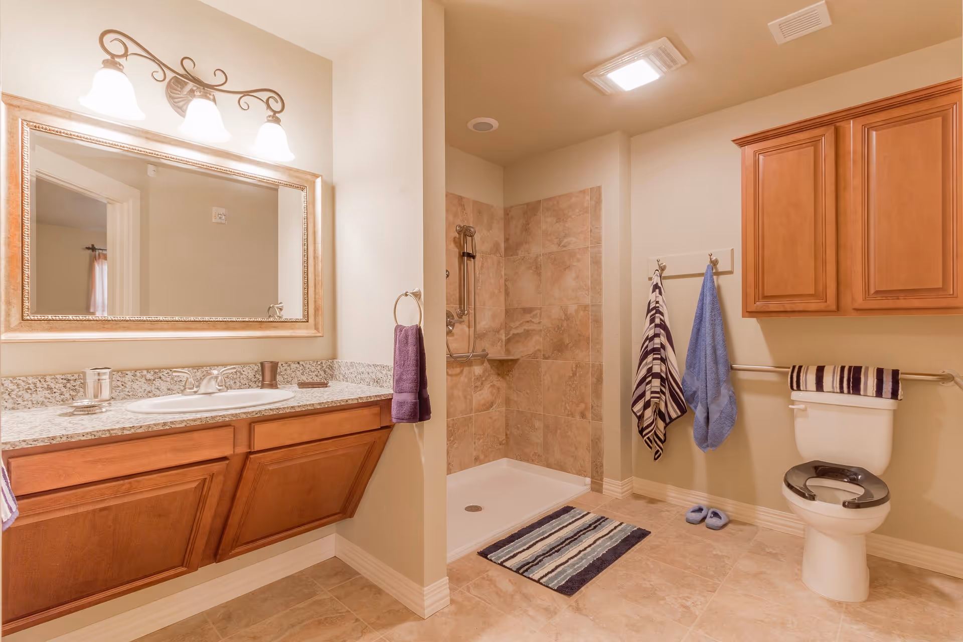 A clean and well-lit bathroom featuring a large mirror above a granite countertop with a sink, wooden cabinets below, a walk-in tiled shower with a grab bar, a toilet with a raised seat and grab bar, striped and solid color towels hanging on hooks and towel bars, and a pair of slippers on the tiled floor.