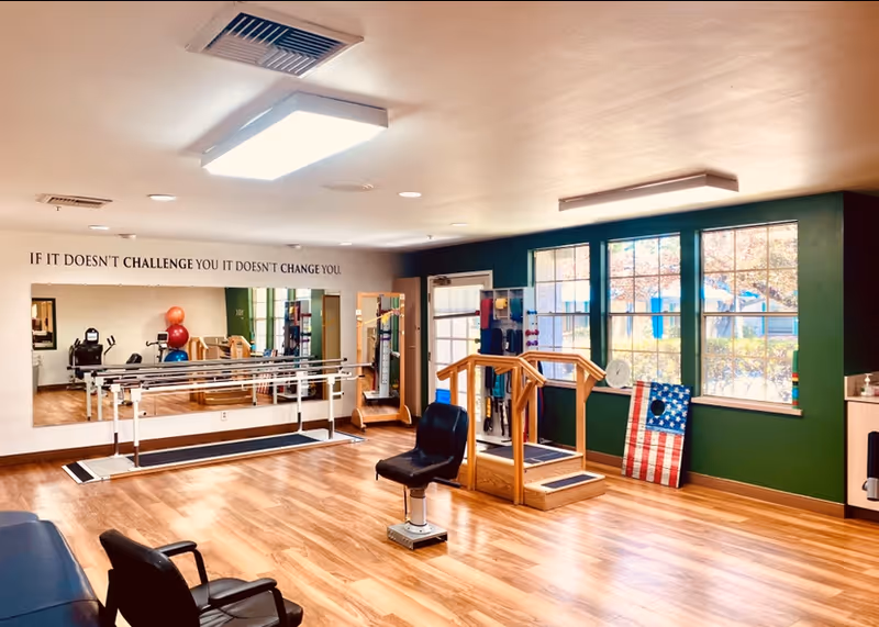 A bright rehabilitation or physical therapy room with wooden flooring, large windows letting in natural light, parallel bars for walking exercises, a small set of stairs with handrails, exercise balls, and a chair in the center. A motivational quote on the wall reads, 'IF IT DOESN'T CHALLENGE YOU IT DOESN'T CHANGE YOU.'