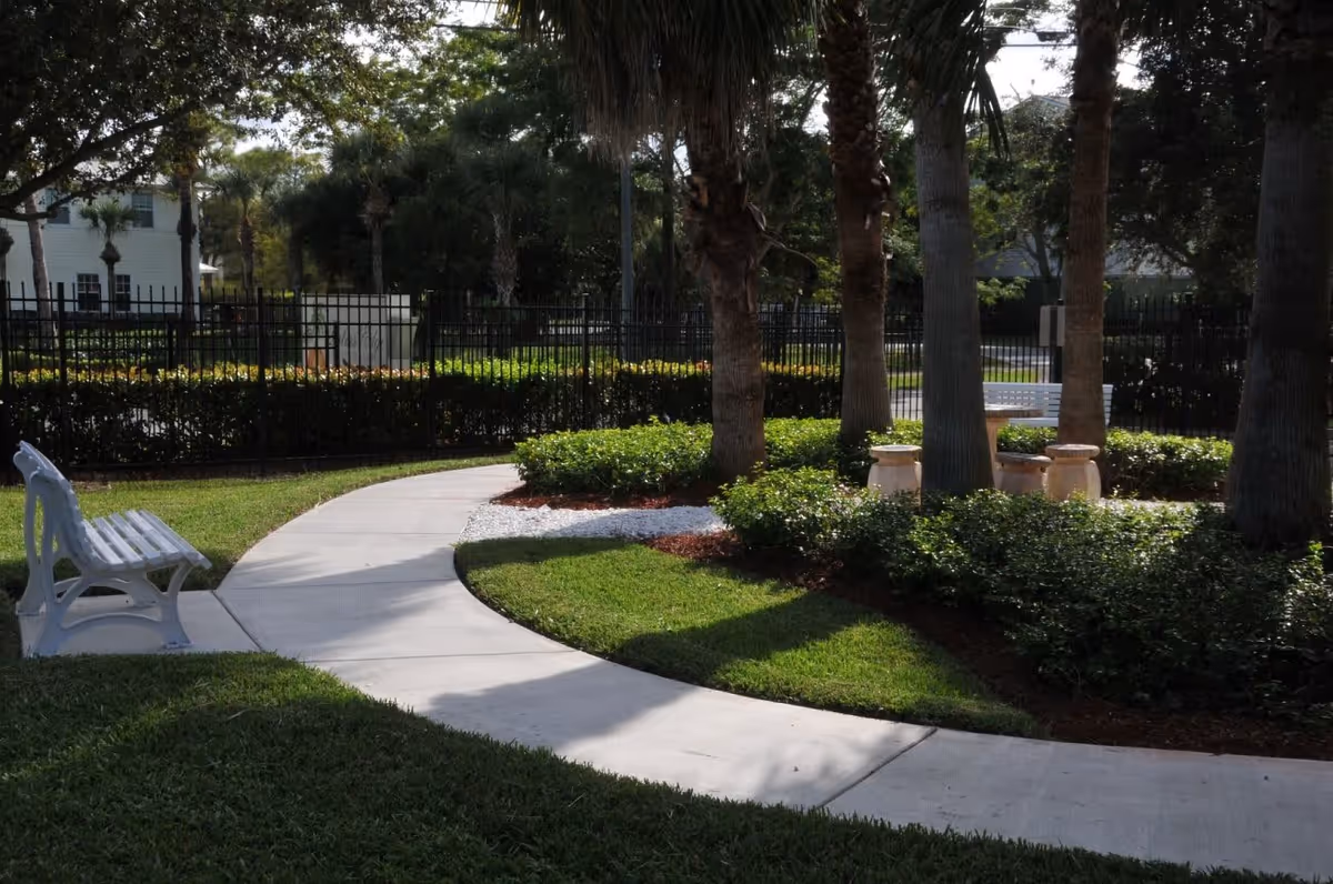 A curved concrete pathway in a garden area with green grass, bushes, and several tall palm trees. There are white benches along the path and a circular seating area with stone stools surrounded by greenery. A black metal fence and residential buildings are visible in the background.
