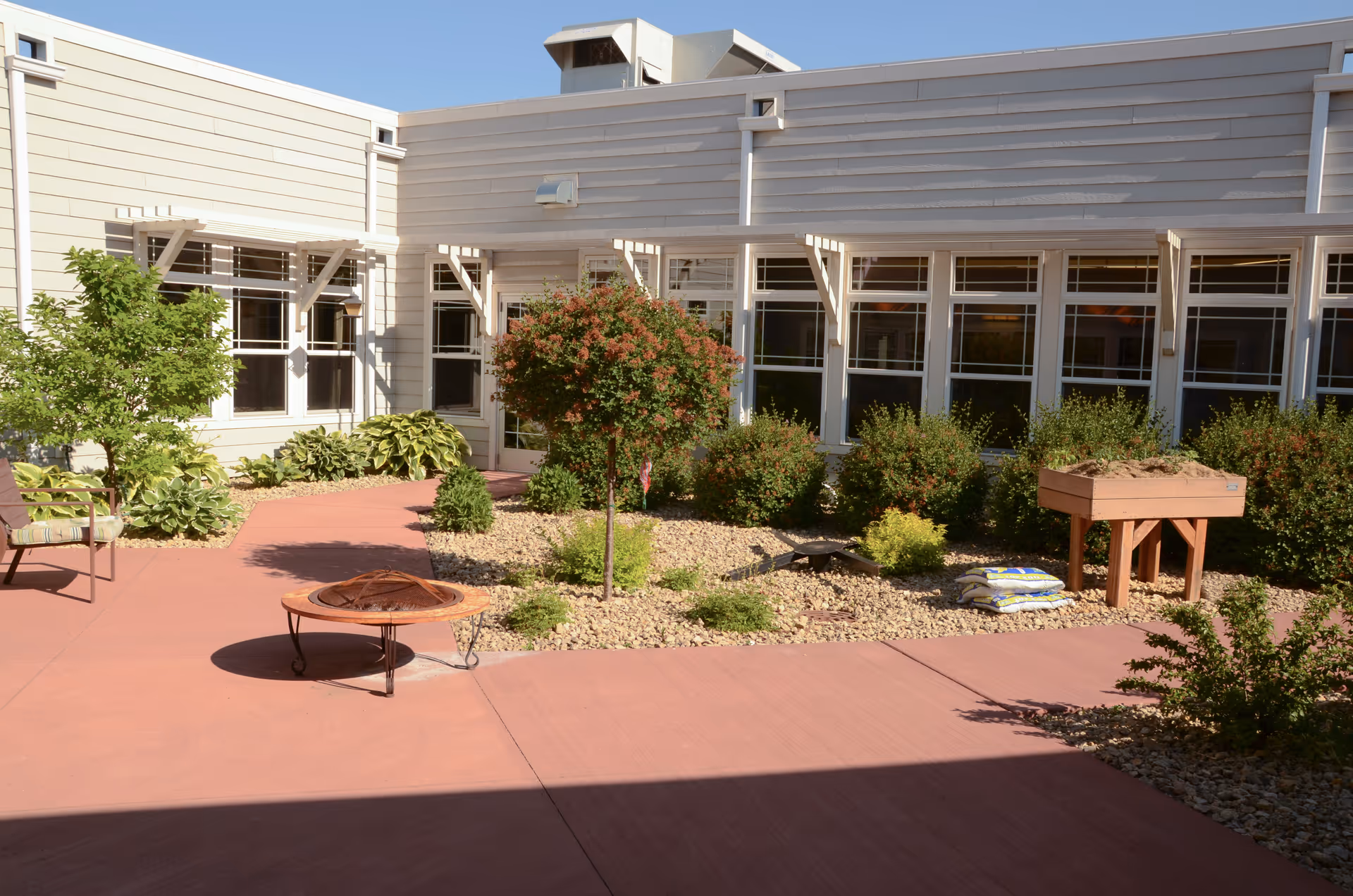 Outdoor courtyard area at Spring Valley Senior Living and Health Care Campus with a paved walkway, small trees and bushes, a fire pit, a chair, and a raised garden bed filled with soil. The building exterior has multiple windows and light-colored siding.