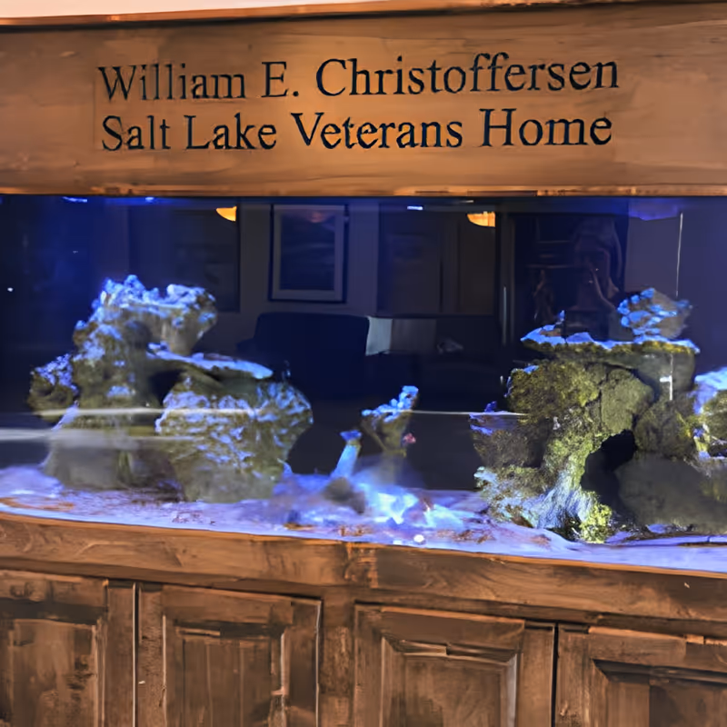 An aquarium with rocks and coral inside a wooden cabinet. Above the aquarium, there is a wooden sign that reads 'William E. Christoffersen Salt Lake Veterans Home'.