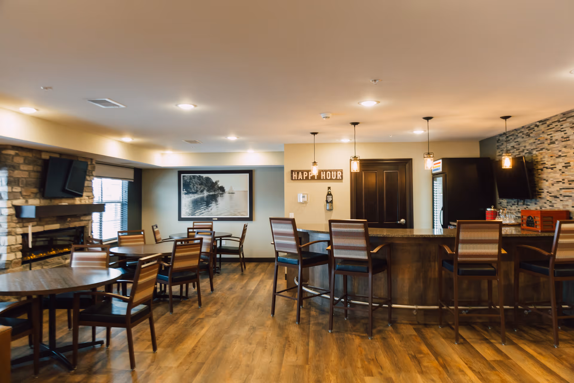 Interior view of a senior living facility common area with wooden floors, several round tables and chairs, a stone fireplace with a mounted TV above it, a bar counter with high chairs, pendant lights hanging from the ceiling, and a wall sign that reads 'HAPPY HOUR'.