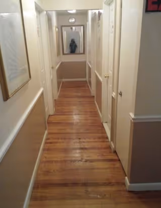 A narrow indoor hallway with wooden flooring and beige walls. There are several white doors on both sides of the hallway, a framed picture on the left wall, and a large mirror at the end reflecting part of the hallway.