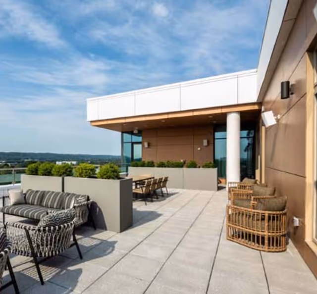 Outdoor patio area with modern seating including a striped cushioned sofa, patterned chairs, and wicker chairs with cushions. There are large planters with green shrubs, a dining table with chairs, and a building wall with windows and a column under a partly cloudy sky.