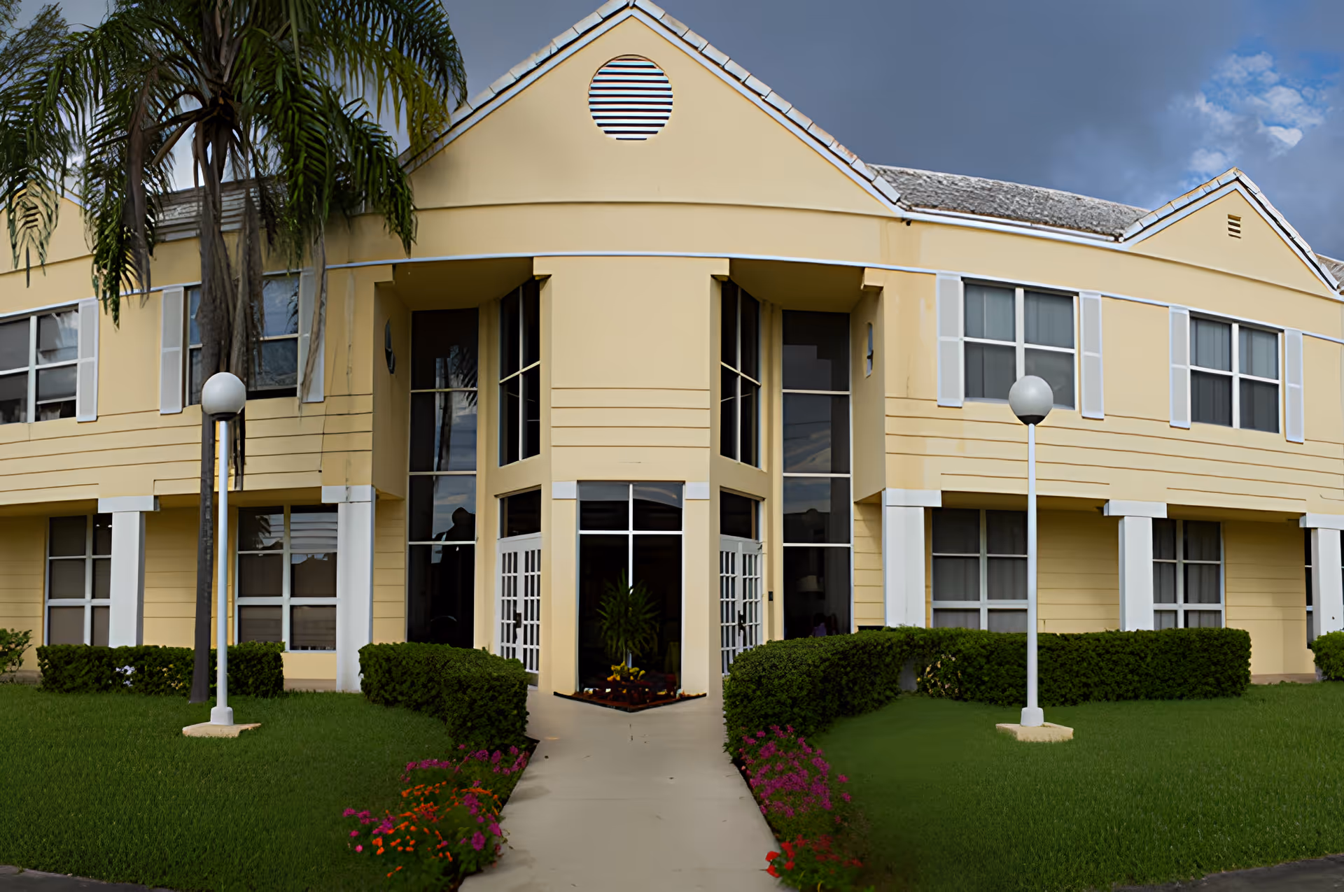Front exterior view of a two-story yellow building with white trim, large windows, and a central entrance. The entrance is flanked by bushes and flower beds, with two lamp posts on either side of the walkway leading to the door. Palm trees are visible on the left side, and the sky is partly cloudy.