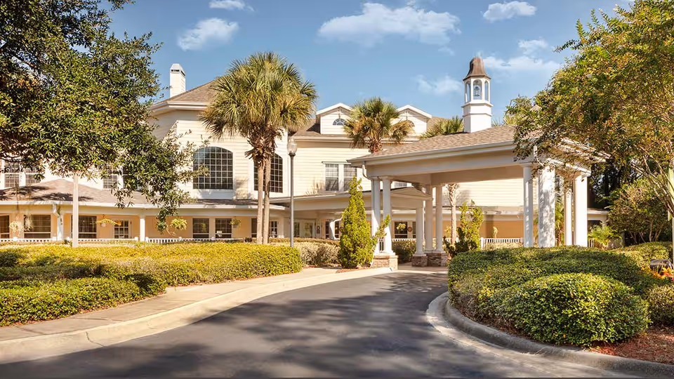 Front entrance and porte-cochère of a two-story light-colored assisted living building with palm trees and landscaped shrubs.