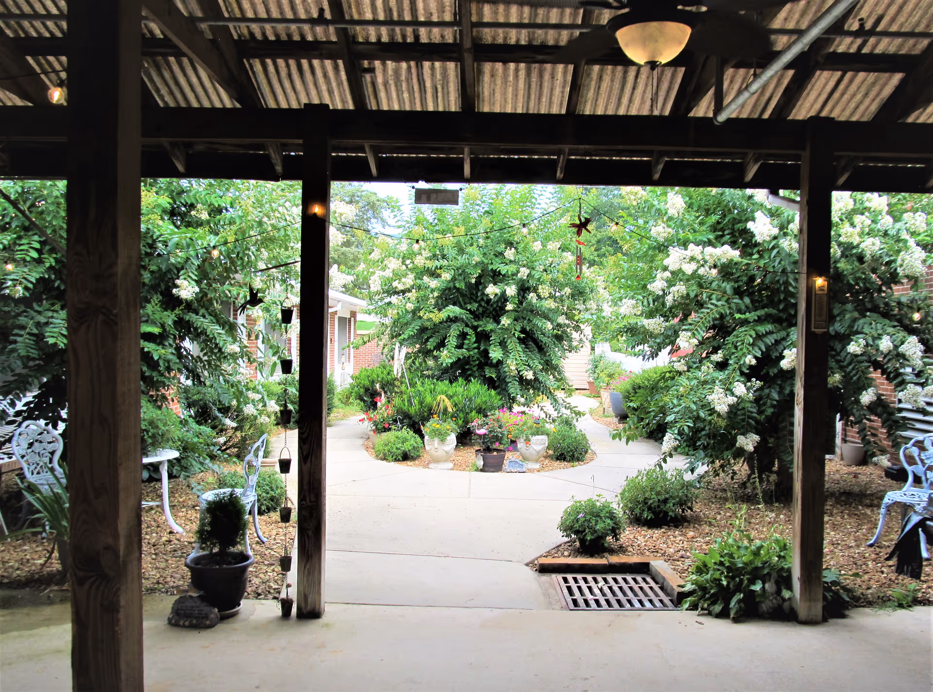 View from a covered patio looking out onto a garden area with lush green bushes, flowering plants, and a concrete pathway. There are white metal chairs and a small table on either side of the patio, with hanging decorations and a ceiling light above.