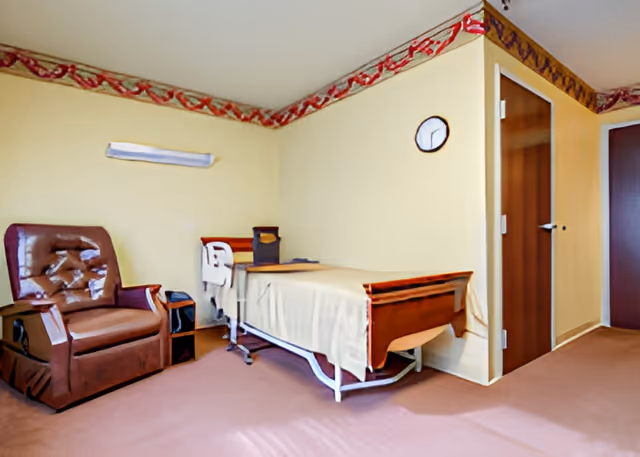 A simple room with a hospital-style bed covered with a beige blanket, a brown recliner chair, a small side table, a wall clock, and a door. The walls are painted light yellow with a decorative border near the ceiling.