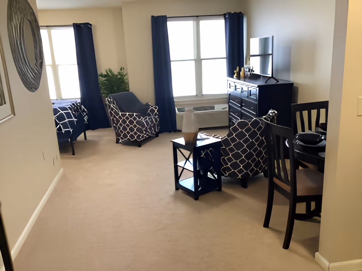 A cozy living area in a senior living facility with beige carpet, two armchairs with patterned cushions, a small side table, a dresser with a TV on top, and a dining table with chairs. Two large windows with dark blue curtains allow natural light into the room. A bed is partially visible in the background.