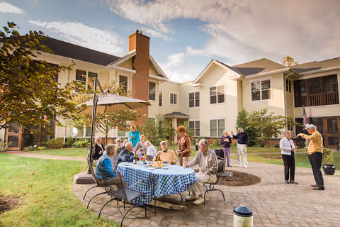 A group of elderly people socializing outdoors in a courtyard of a senior living community. Some are seated around a table with a blue checkered tablecloth, enjoying food and drinks, while others stand and chat nearby. The courtyard is surrounded by two-story residential buildings with white siding and brick chimneys under a partly cloudy sky.