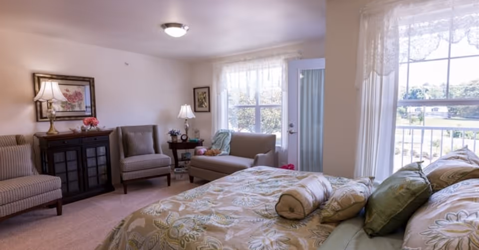 A bright and cozy bedroom in a senior living facility featuring a large bed with floral bedding and multiple pillows. The room includes two armchairs, a small sofa near a window with sheer curtains, a dark wooden cabinet with a lamp and framed artwork above it, and a side table with another lamp and decorative items. A door with a glass panel leads outside, allowing natural light to fill the room.