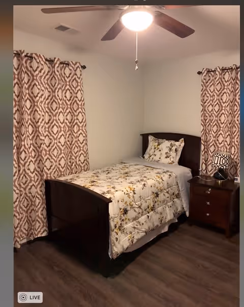 A small bedroom with a single dark wooden bed frame, a floral patterned comforter and matching pillow, two windows with patterned curtains, a ceiling fan with light, and a wooden nightstand with a lamp and a small clock.