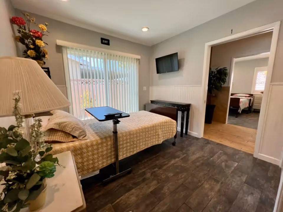 Small furnished bedroom with a single bed, overbed table, wall-mounted TV, sliding vertical blinds and an open doorway to another room.