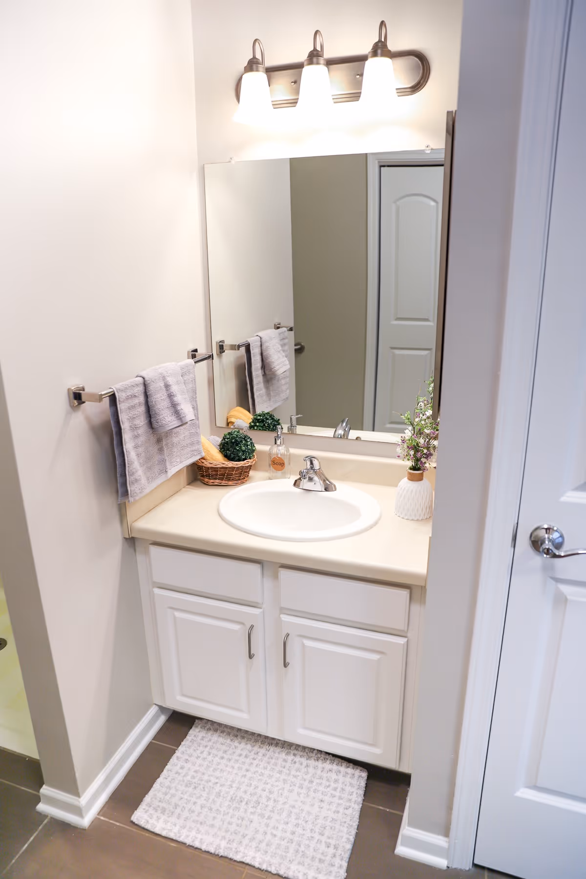 A clean and well-lit bathroom vanity area with a white sink, beige countertop, and white cabinets underneath. Above the sink is a large mirror with three light fixtures mounted on the wall. A gray towel hangs on a towel rack to the left, and decorative items including a small basket with greenery and a soap dispenser are placed on the countertop. A white textured rug is on the floor in front of the vanity.