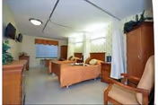 Two hospital-style beds in a nursing home room with chairs, a TV, cabinets, and privacy curtains.