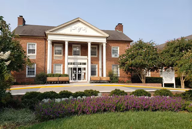 Front exterior view of a two-story brick building with white columns at the entrance, benches on either side of the door, and well-maintained landscaping including purple flowers and green shrubs in the foreground.