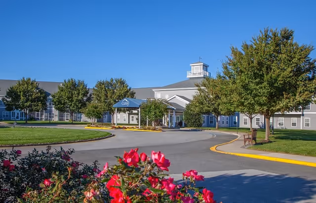 Exterior view of a senior living facility with a circular driveway, green lawns, trees, and blooming pink flowers in the foreground under a clear blue sky.