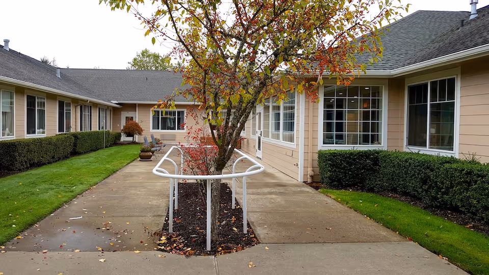 Outdoor courtyard area of a senior living facility with a paved walkway, a tree surrounded by a white metal railing, green grass, trimmed bushes, and beige buildings with multiple windows on either side.