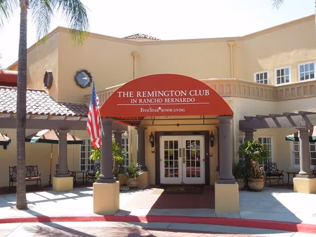 Front entrance of The Remington Club senior living facility with a red awning, double doors, columns, potted plants and an American flag.