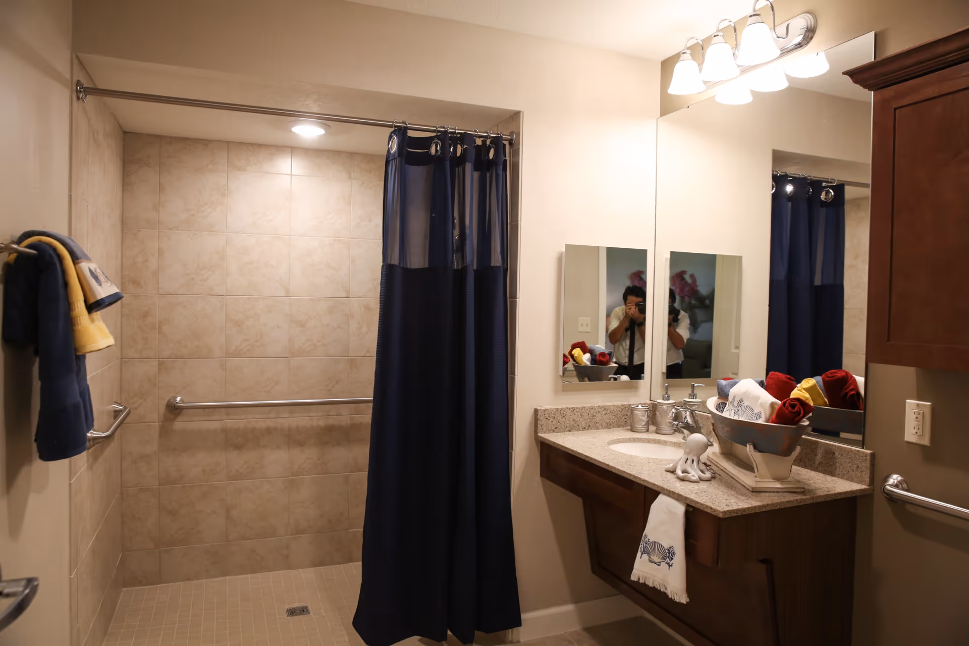 A bathroom with a tiled walk-in shower featuring a navy blue shower curtain. There are grab bars inside the shower and on the wall near the sink. The vanity has a granite countertop with a sink, a decorative bowl holding rolled towels in red, yellow, and white, and a small octopus figurine. Above the vanity is a large mirror with a light fixture containing four bulbs. Towels hang on a rack to the left of the shower.
