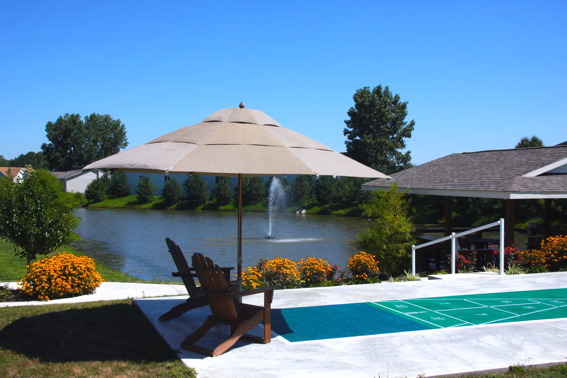 Outdoor seating area with two wooden chairs under a large beige umbrella next to a shuffleboard court. In the background, there is a pond with a water fountain, surrounded by green trees and colorful flowers under a clear blue sky.