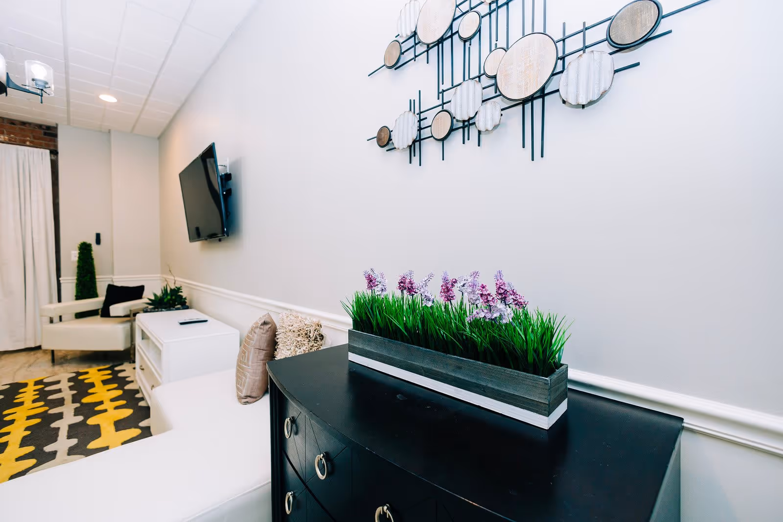 A modern living room with a black console table topped with a rectangular planter of green grass and purple flowers. On the wall above the table is a decorative metal wall art featuring circular shapes. In the background, there is a white armchair with a black pillow, a white media console with a TV mounted on the wall above it, and a patterned rug with yellow, gray, and white colors.