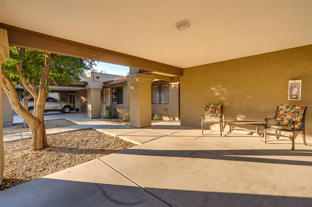 Covered outdoor patio area with two cushioned chairs and a small table, adjacent to a building with beige walls and windows. A tree and gravel landscaping are visible on the left side, along with a parked car in the background.