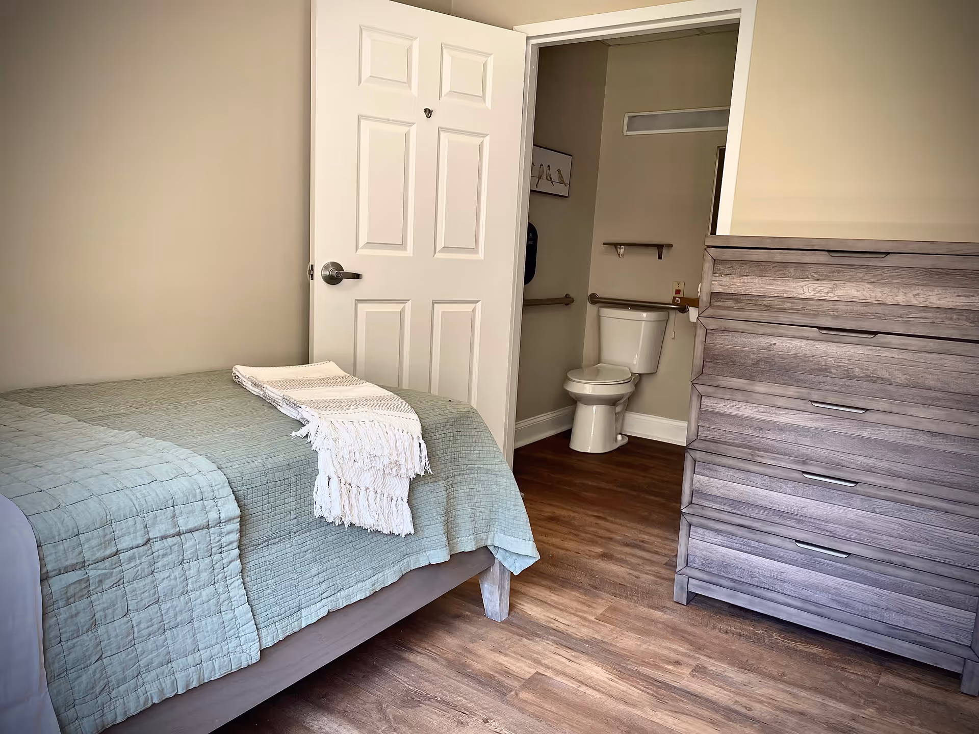 Small bedroom with a bed, wooden dresser, and an open door revealing an attached bathroom with a toilet.