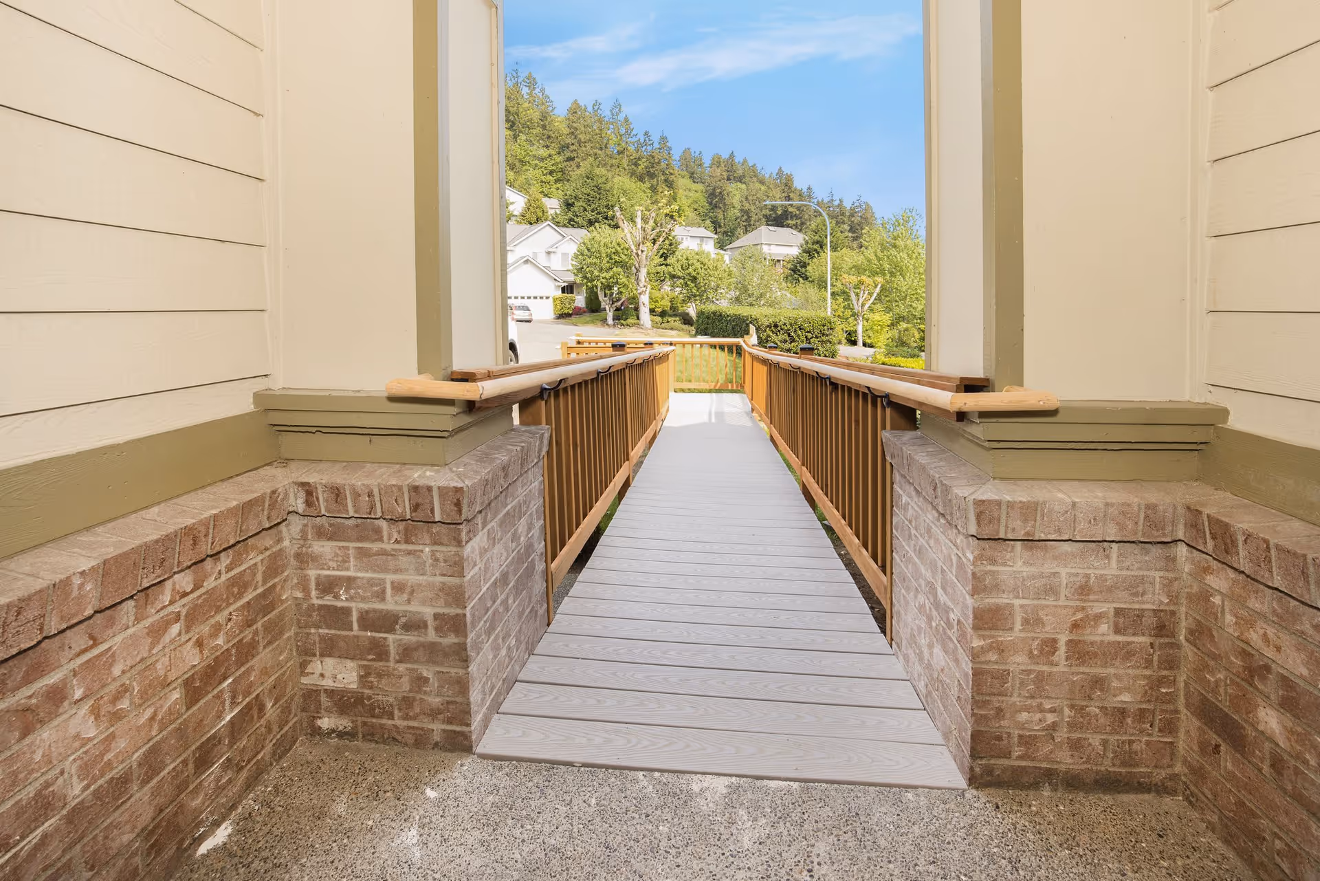 A wheelchair accessible ramp with wooden handrails on both sides leading from a building entrance to an outdoor area with trees and houses in the background under a clear blue sky.