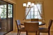 A dining area with a round wooden table surrounded by four upholstered chairs. A chandelier with five lights hangs above the table. Sheer curtains are drawn to the sides of a window, and a glass door leads outside, allowing natural light to fill the room.