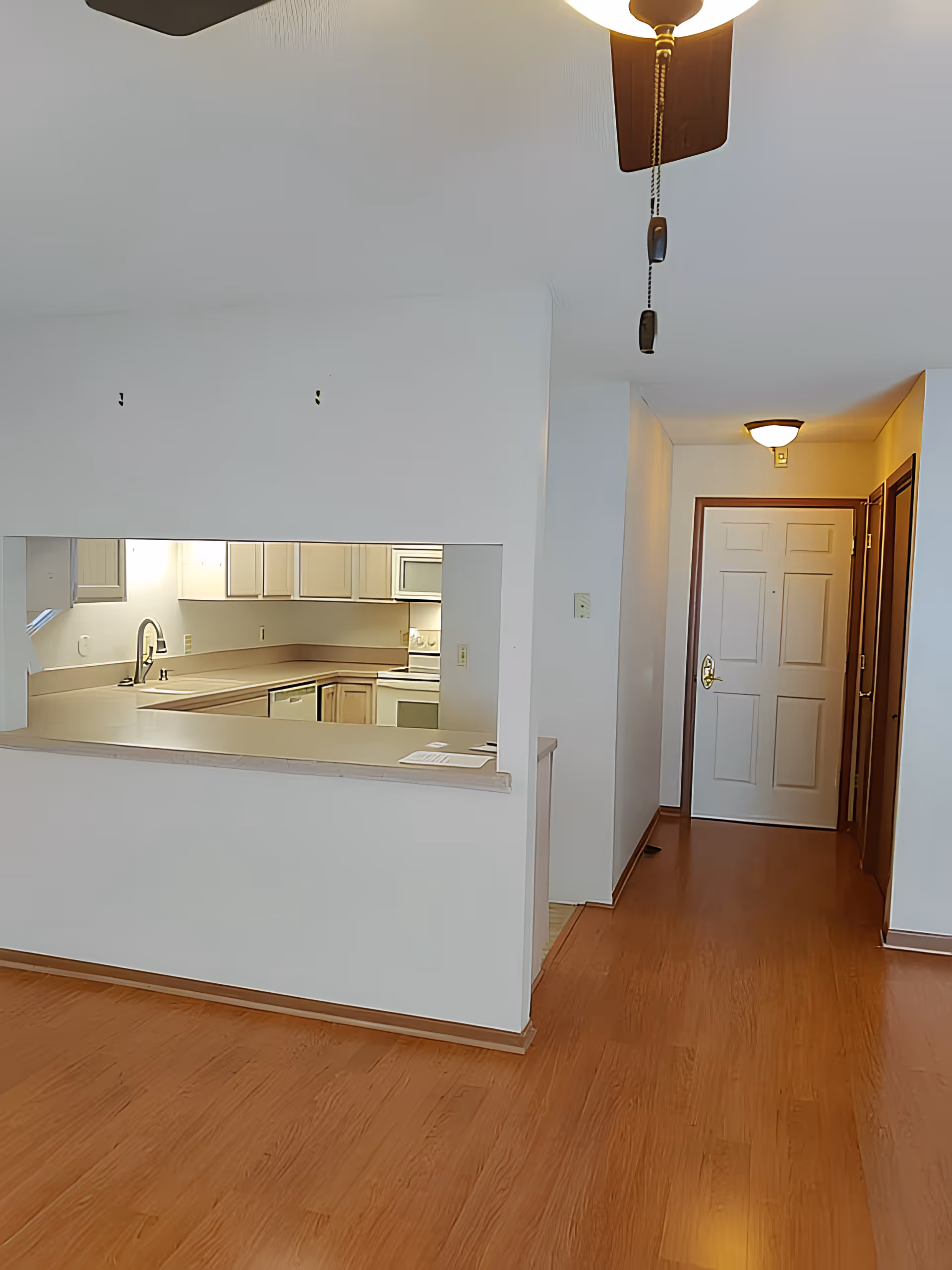 Interior view of a residential space showing a kitchen with white cabinets and appliances, a countertop with a pass-through window, a hallway with a white door at the end, wooden flooring, and ceiling lights including a ceiling fan with pull chains.