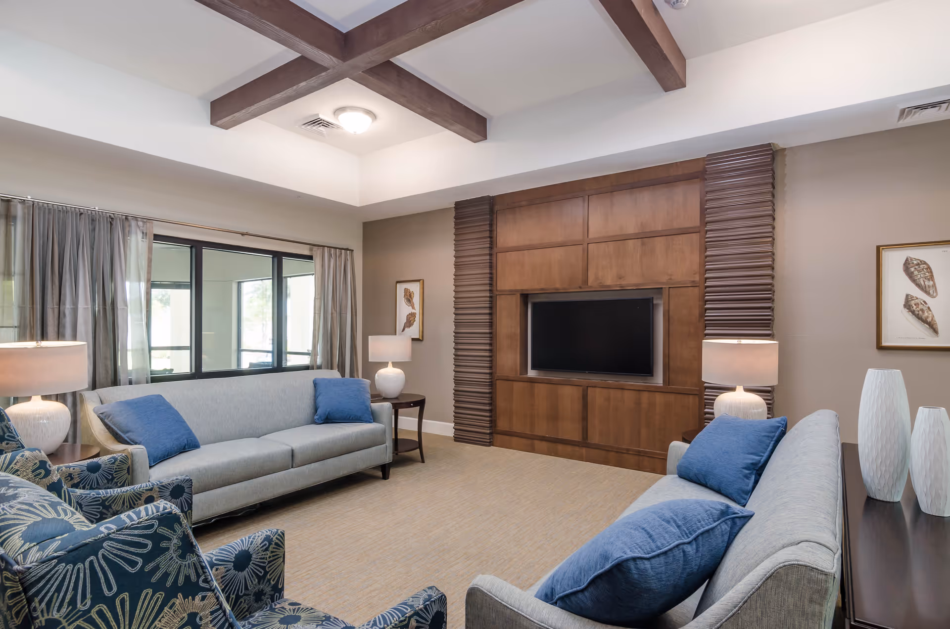 A cozy living room with two light gray sofas adorned with blue pillows, two patterned armchairs, a wooden entertainment center with a flat-screen TV, two table lamps on side tables, and decorative vases. The room features a beige carpet, large windows with curtains, and a ceiling with wooden beams.