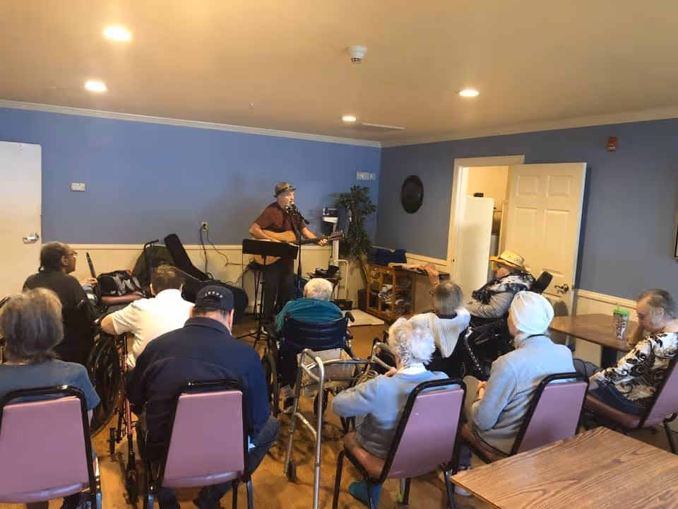 A group of elderly people seated in chairs and wheelchairs facing a man playing guitar and singing in a room with blue walls and wooden floor. The room appears to be a common area or lounge in an assisted living facility.