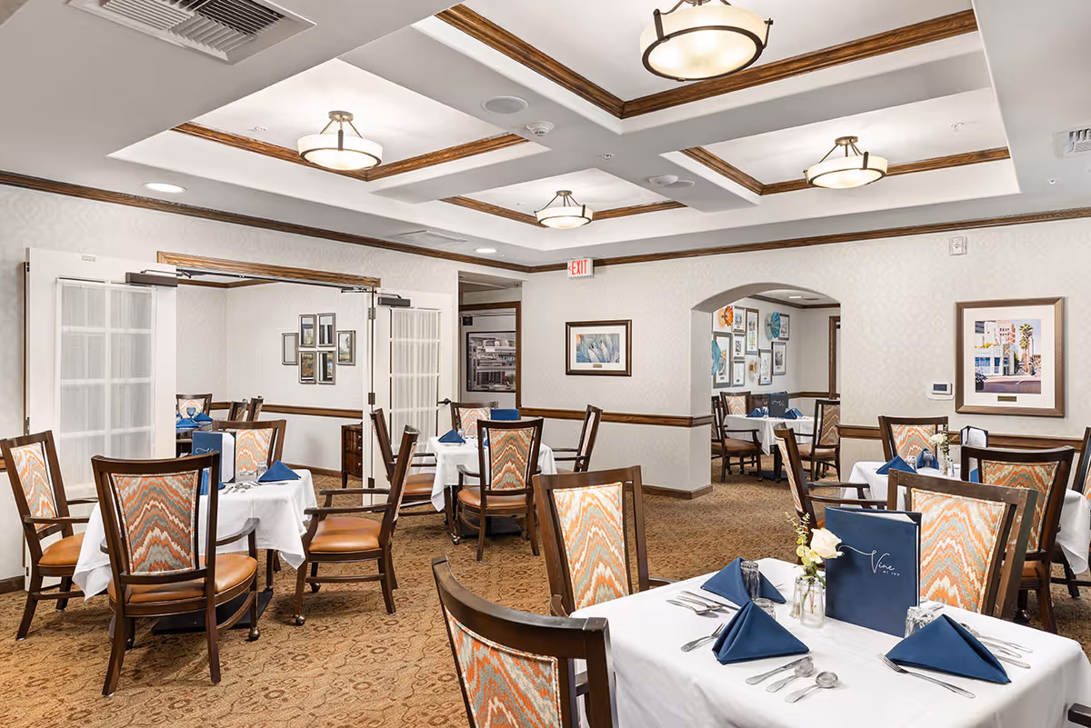 A well-lit dining room with multiple tables covered in white tablecloths, each set with blue napkins, silverware, and small flower vases. The room features patterned carpet, wooden chairs with colorful upholstery, framed artwork on the walls, and ceiling lights with wooden accents.