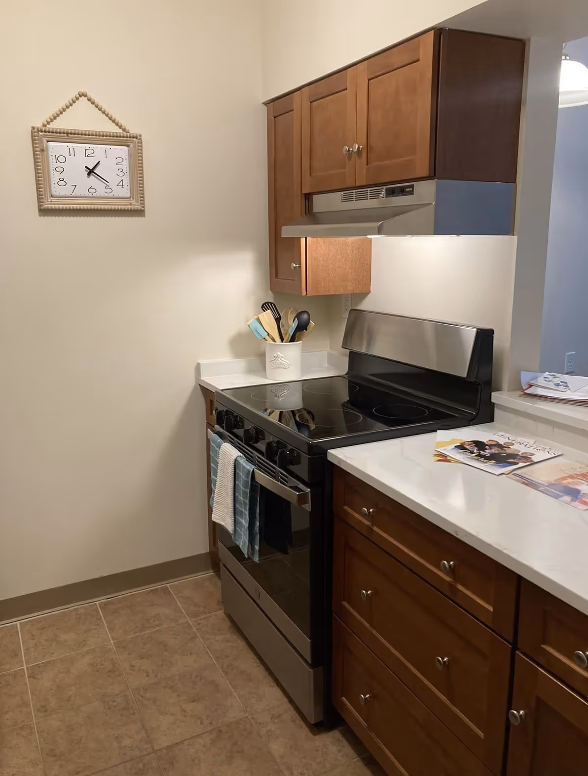 A kitchen corner with a stainless steel electric stove and oven, wooden cabinets above and below the countertop, a utensil holder with cooking tools on the counter, a wall clock showing 10:25, and some magazines on the countertop.