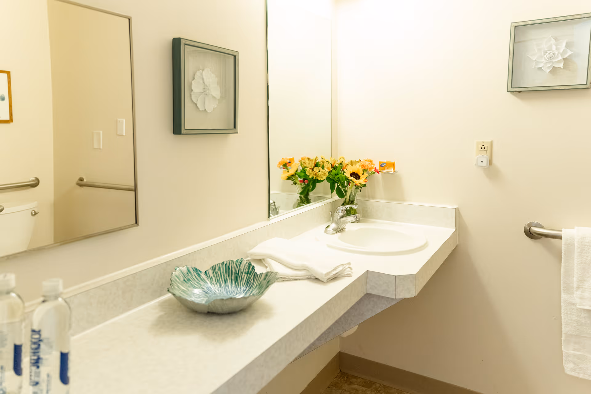 A clean bathroom sink area with a large mirror above the countertop. On the counter, there is a decorative green bowl, two bottles of water, a folded white towel, and a small vase with yellow and orange flowers. The walls are light-colored, and there is a towel hanging on a grab bar to the right. Two framed floral artworks are mounted on the walls.