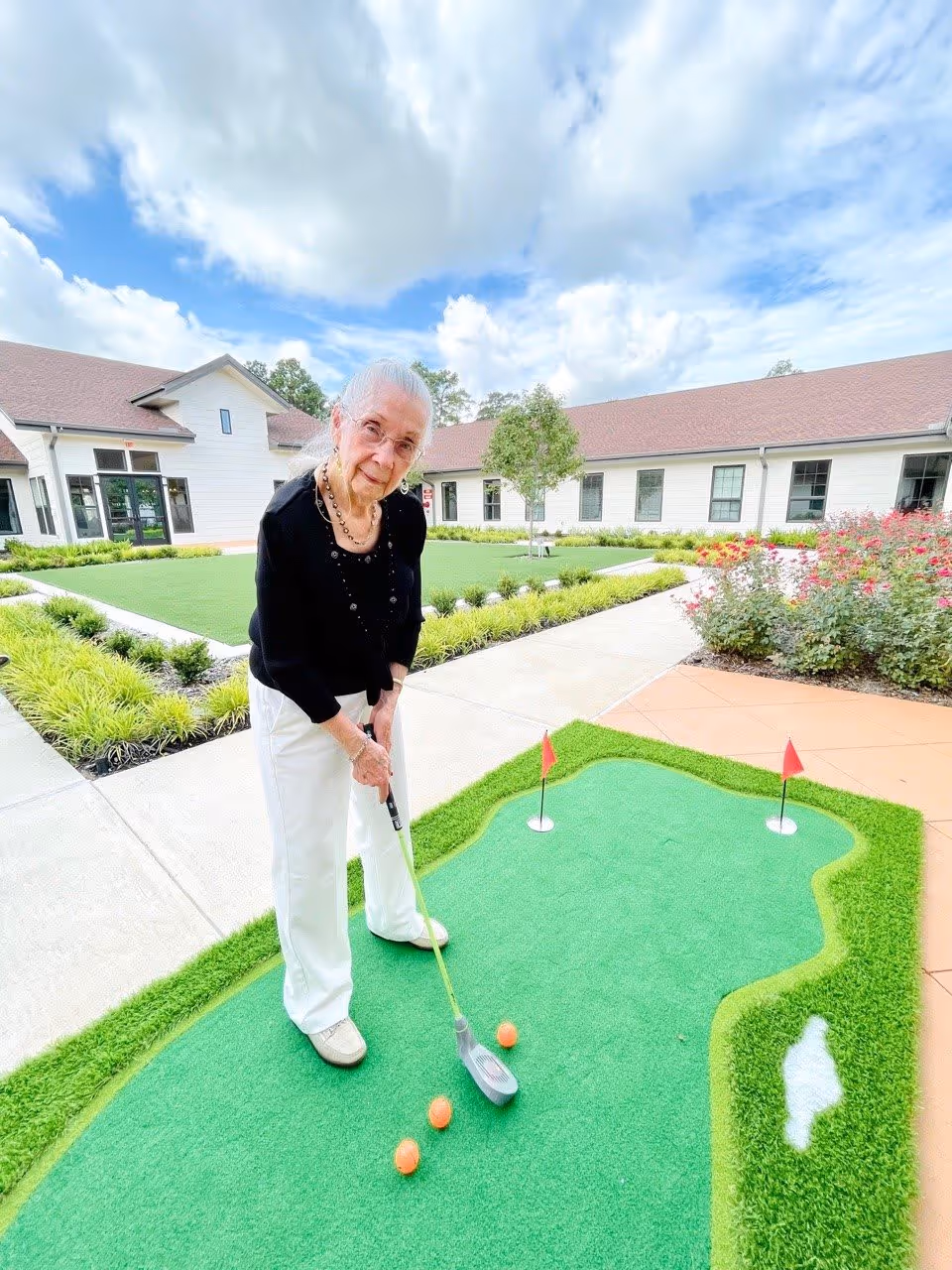 An elderly woman with white hair, glasses, and a black top is playing mini golf on a small putting green with three orange golf balls. She is standing outdoors in a courtyard area with a building in the background, green grass, bushes, and a partly cloudy sky.