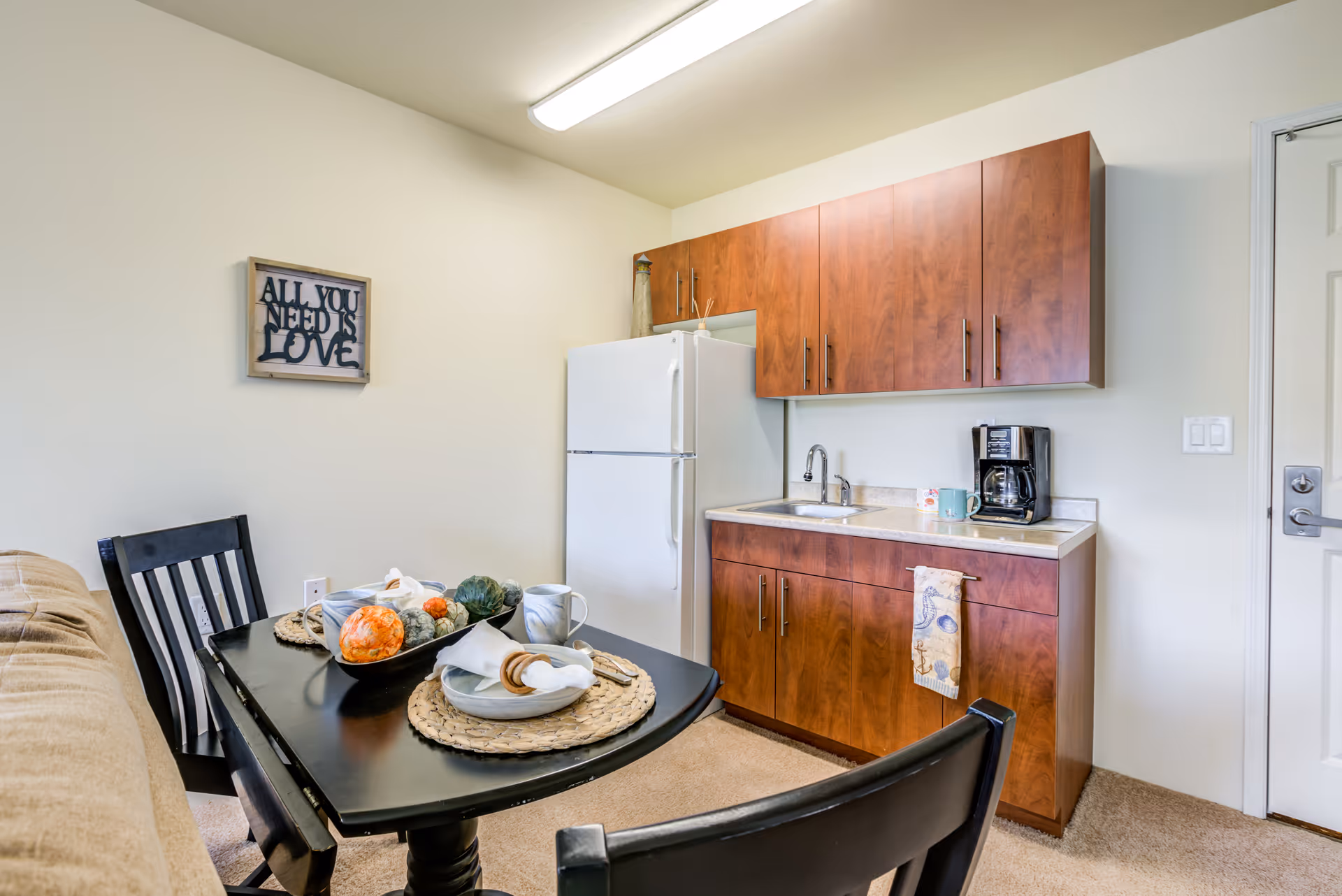 A small kitchen area with wooden cabinets, a white refrigerator, a sink, and a coffee maker on the counter. In front of the kitchen is a black dining table set with two place settings, decorative pumpkins, and two black chairs. A beige couch is partially visible on the left side. A wall decoration with the text 'ALL YOU NEED IS LOVE' hangs on the wall above the dining table.