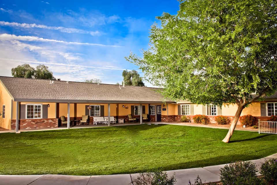 Single-story senior living facility building with a covered porch featuring outdoor seating, surrounded by a well-maintained green lawn and a large leafy tree under a partly cloudy blue sky.
