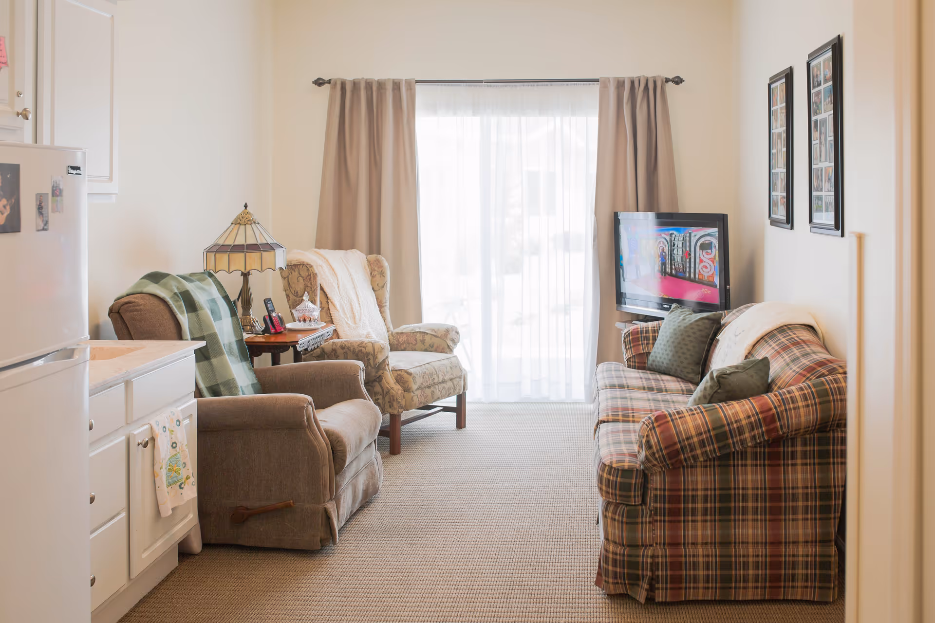 A cozy living room area with a plaid sofa, two armchairs, a side table with a lamp and phone, a wall-mounted TV, and large windows covered with sheer curtains and beige drapes letting in natural light.