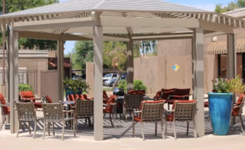 Outdoor seating area with multiple chairs and tables under a beige pergola structure, surrounded by potted plants and buildings in the background.